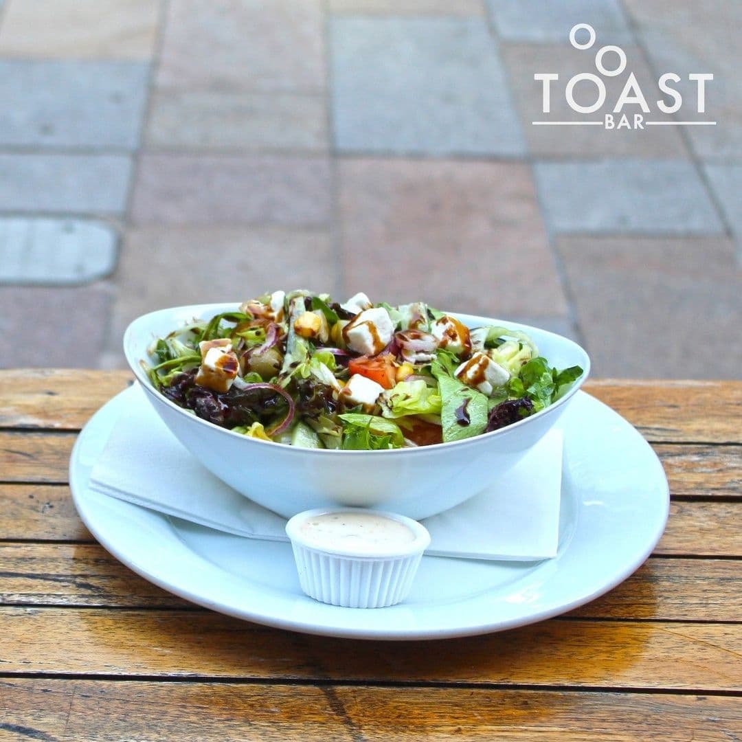 Fresh mixed green salad with assorted vegetables and feta cheese, served with a side of dressing at Toast Bar.
