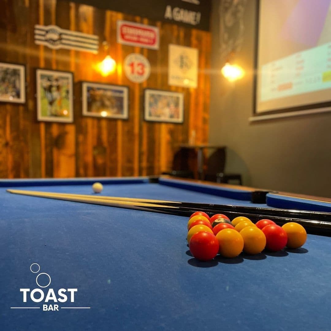A close-up view of a blue billiard table with a triangle of red and yellow pool balls, cue sticks, and a white cue ball at Toast Bar, with a warm, wood-paneled wall adorned with various signs and framed pictures in the background.