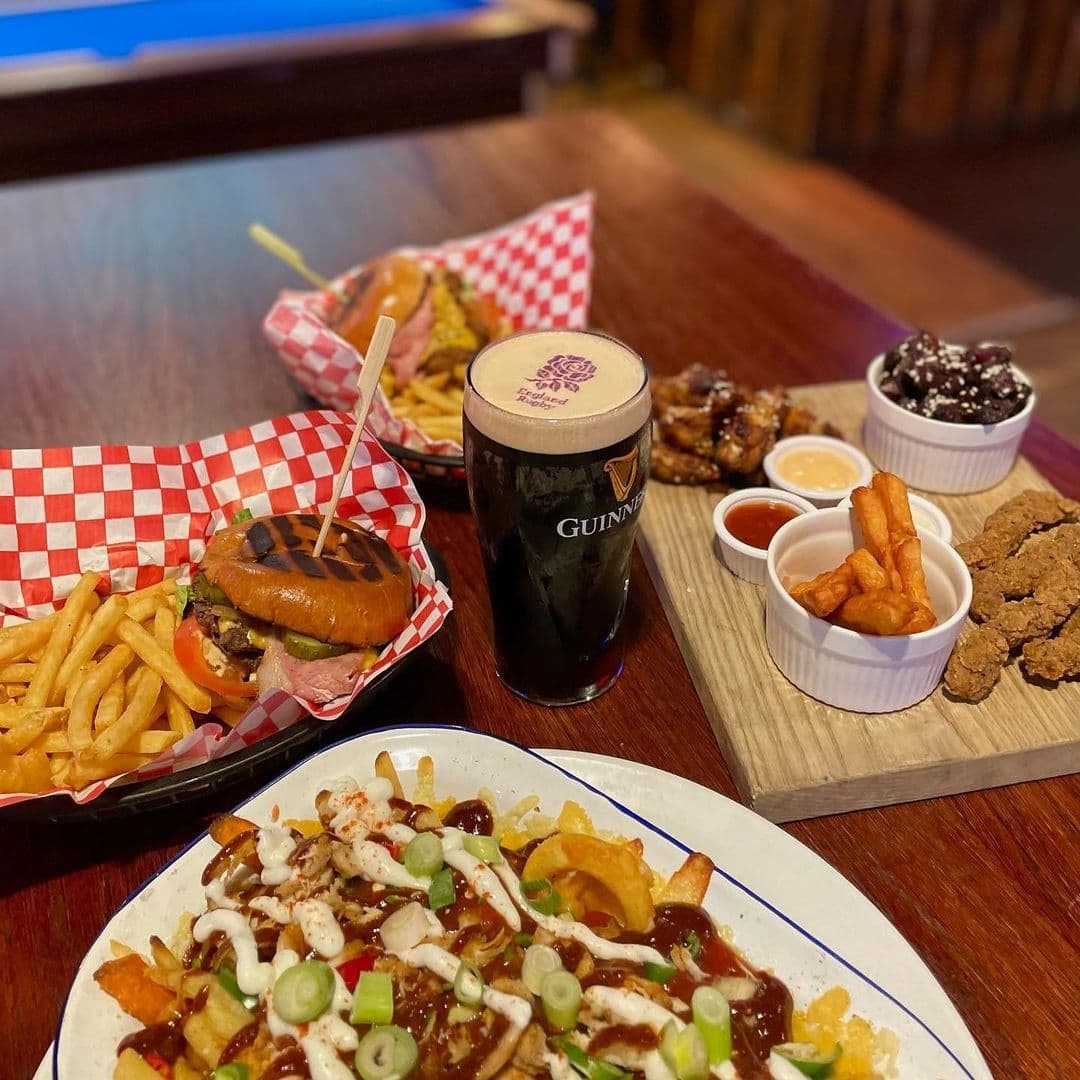 A variety of pub food and a pint of Guinness beer on a wooden table at Toast Bar, featuring a cheeseburger with fries, chicken wings, mozzarella sticks, and loaded fries.