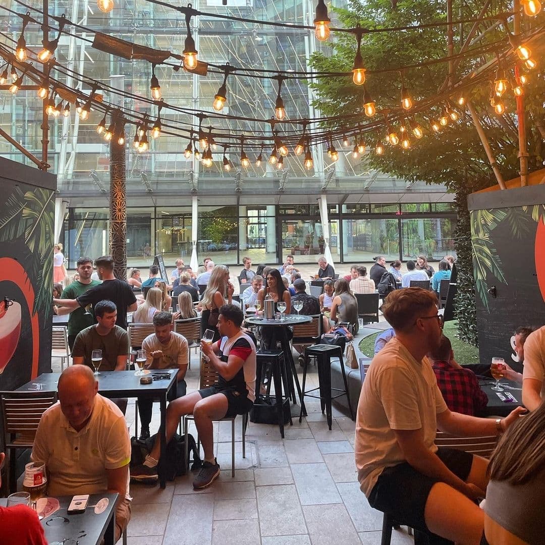 Outdoor seating area at Toast Bar with patrons enjoying drinks under string lights, surrounded by greenery and modern architecture.