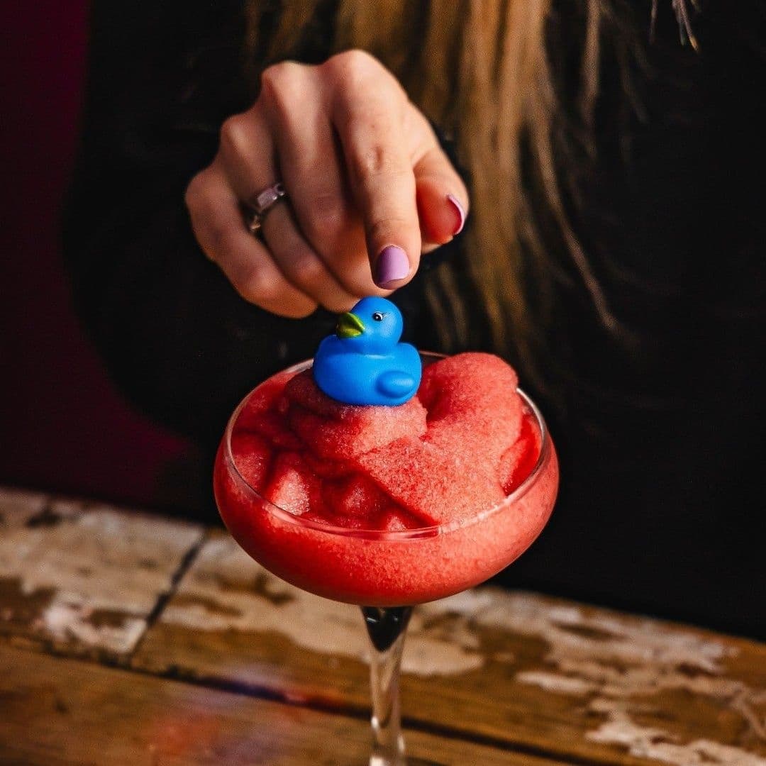 Hand holding a vibrant red cocktail with a blue rubber duck garnish at Cuckoo.