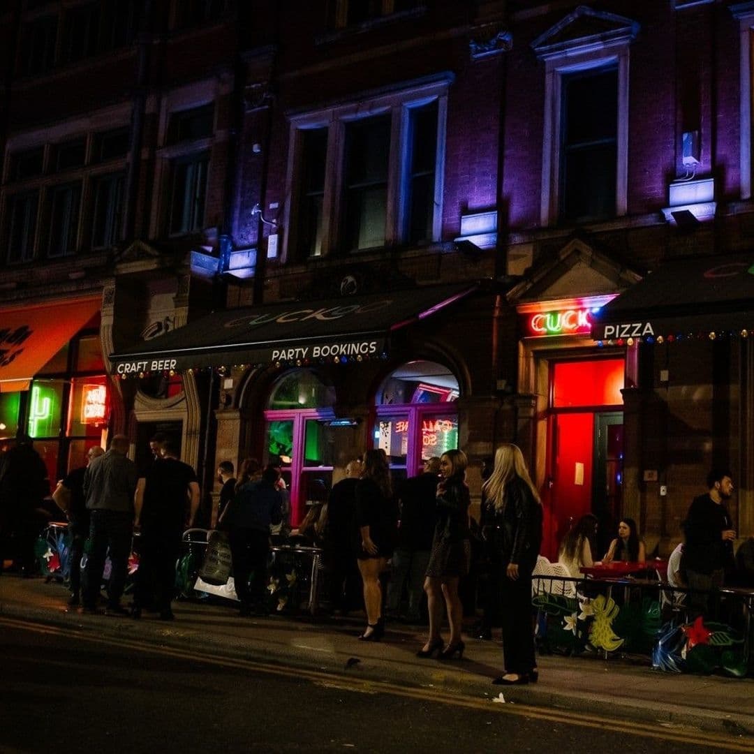 Nighttime view of lively street scene outside Cuckoo, with people gathered and neon signs advertising craft beer and pizza.