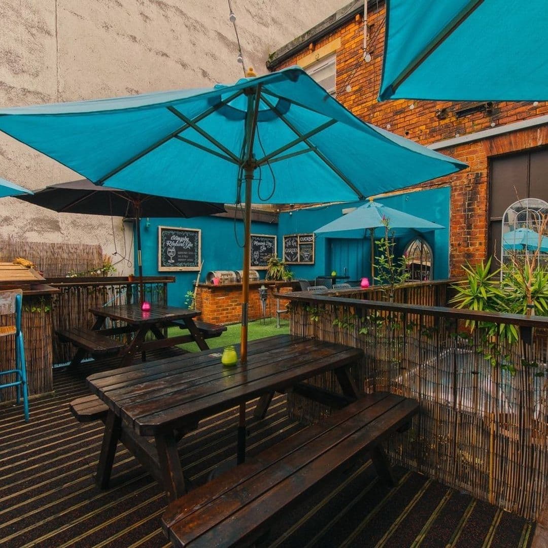 Outdoor seating area at Cuckoo with wooden picnic tables, blue umbrellas, and green plants against a backdrop of brick walls with chalkboard menus.