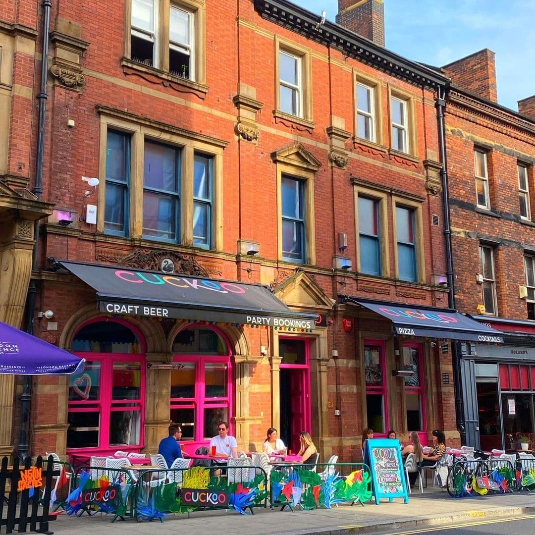 Exterior view of Cuckoo, featuring patrons seated at outdoor tables with colorful barriers, under a sign advertising craft beer and party bookings, set against a traditional red-brick building.