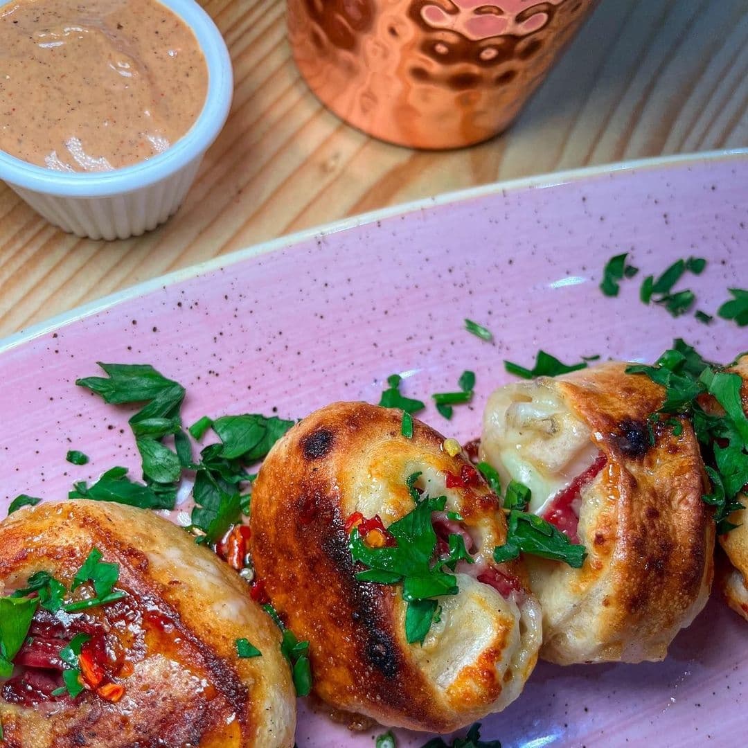 Freshly baked garlic knots topped with melted cheese and parsley, served with a side of spicy dipping sauce on a pink plate at Ecco Pizzeria.