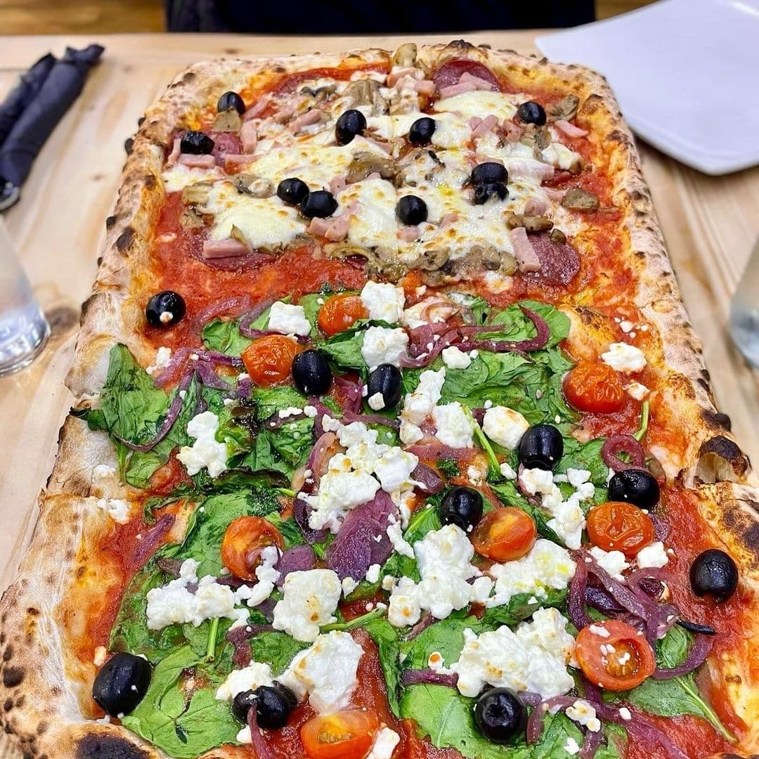 Rectangular pizza with various toppings on a wooden table at Ecco Pizzeria. The pizza is topped with tomato sauce, melted cheese, spinach, black olives, cherry tomatoes, red onions, and chunks of feta cheese, with a golden-brown crust visible on the edges.