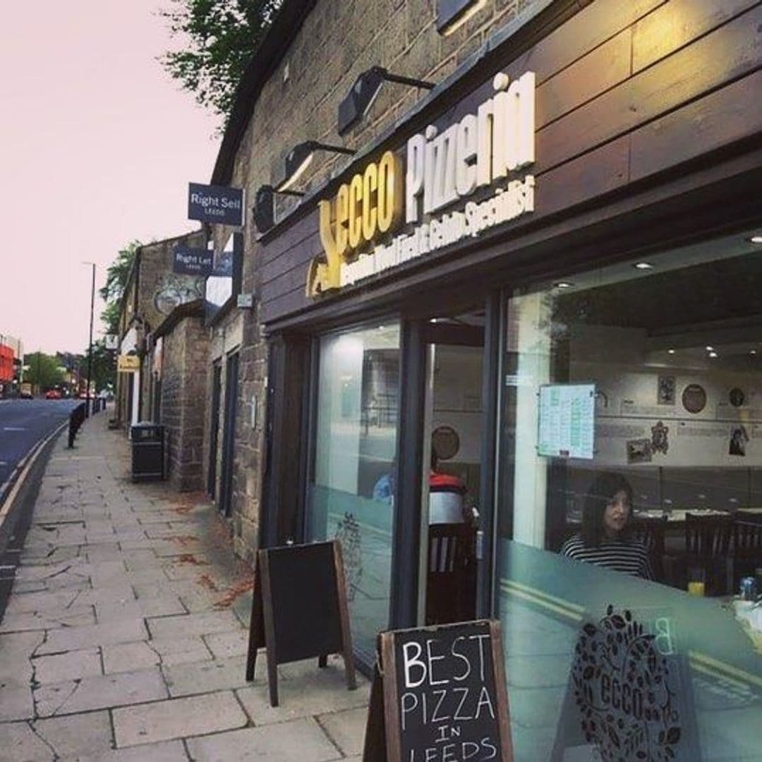Exterior view of Ecco Pizzeria with signage, sidewalk with a chalkboard sign reading "BEST PIZZA IN LEEDS," and a glimpse of the interior seating area.