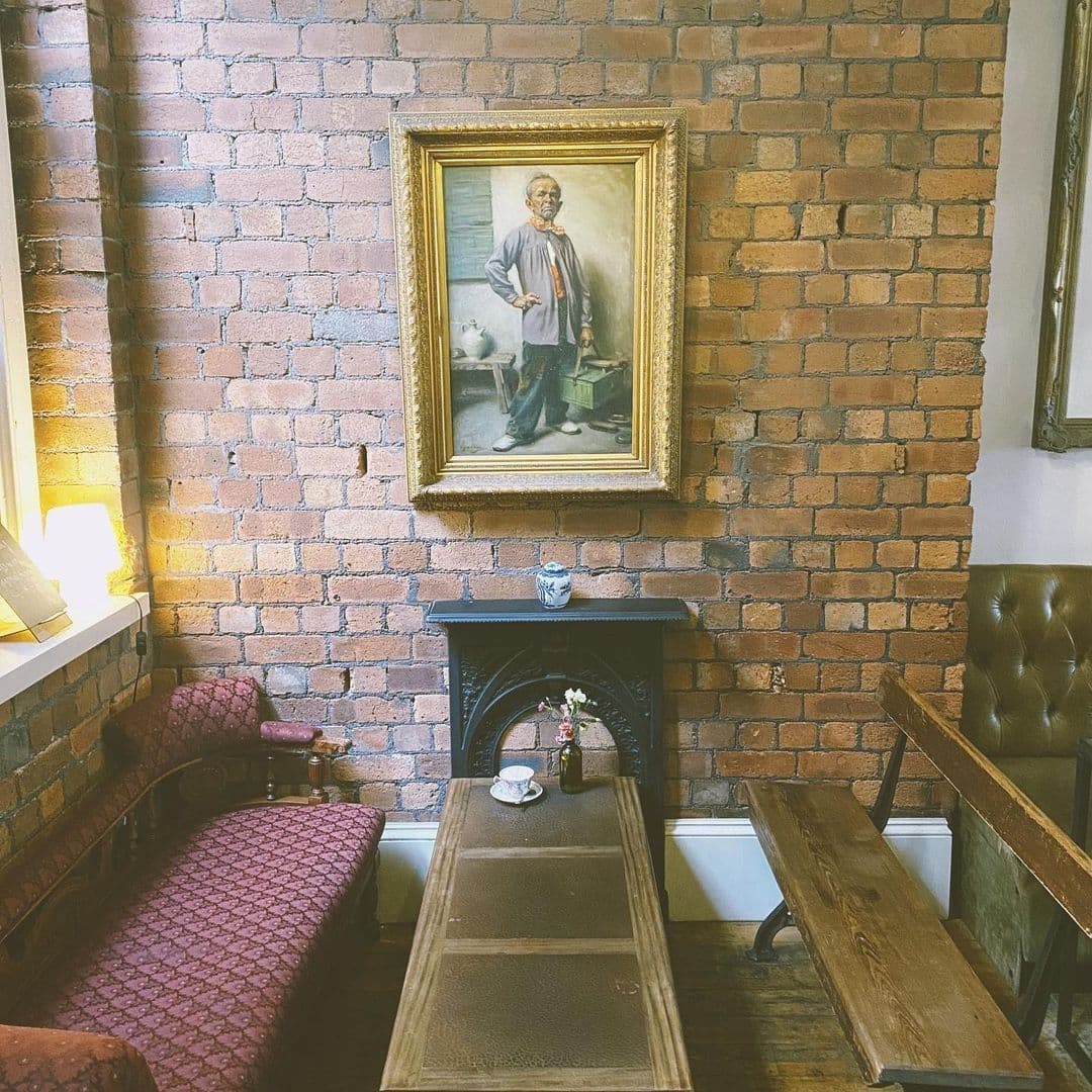 Cozy corner at Mrs Atha's with exposed brick walls, vintage portrait painting, and classic fireplace, complemented by patterned seating and wooden furniture.