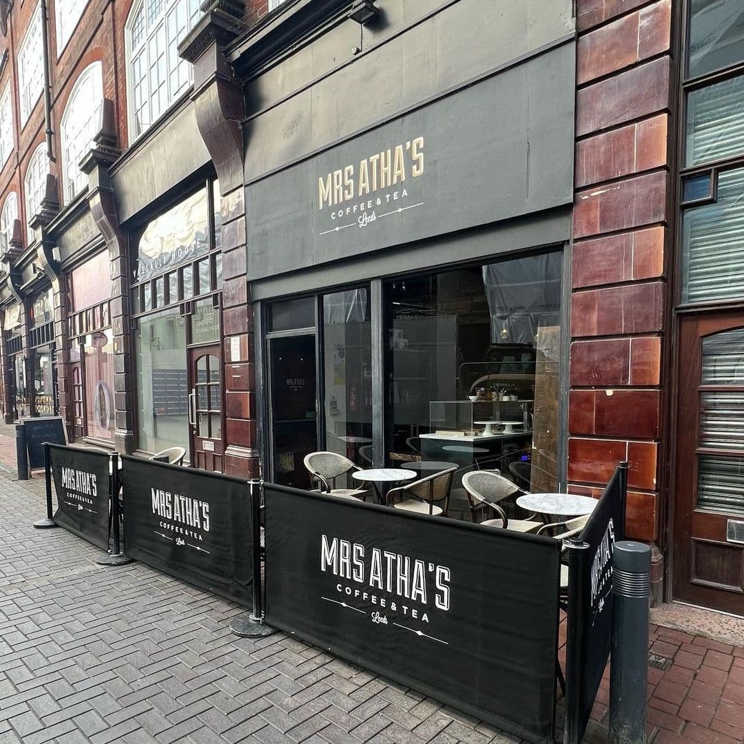 Exterior view of Mrs Atha's coffee and tea shop with outdoor seating behind black barriers featuring the cafe's name, set against a traditional brick building façade.