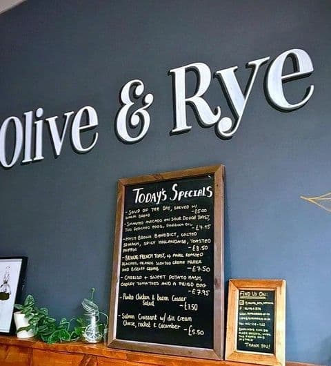 Interior wall of Olive & Rye featuring the restaurant's name in large, stylish letters above a chalkboard displaying the day's specials, with decorative plants on a shelf.