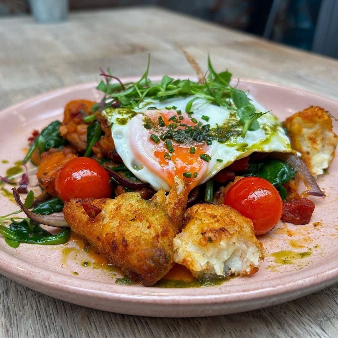 Gourmet brunch dish at Olive & Rye featuring a poached egg with runny yolk, crispy fried cauliflower, fresh spinach, cherry tomatoes, and a drizzle of pesto on a pink ceramic plate.