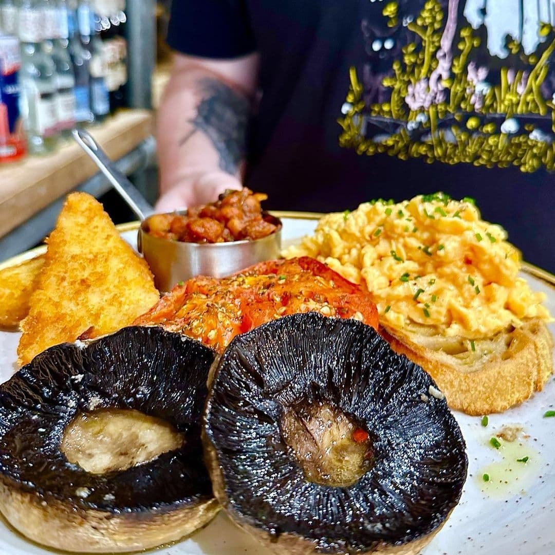 Alt text: "Gourmet breakfast served at Olive & Rye featuring scrambled eggs, grilled tomatoes, hash browns, baked beans, and portobello mushrooms on a white plate, presented by a person with a tattooed arm."