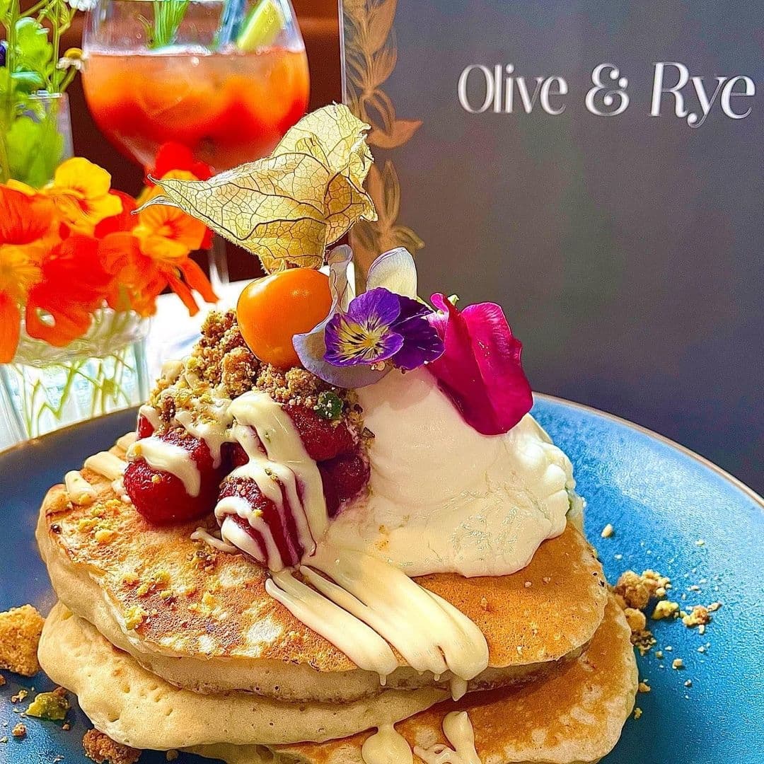 Stack of pancakes with fresh berries, cream, and edible flowers at Olive & Rye, with a refreshing cocktail in the background.