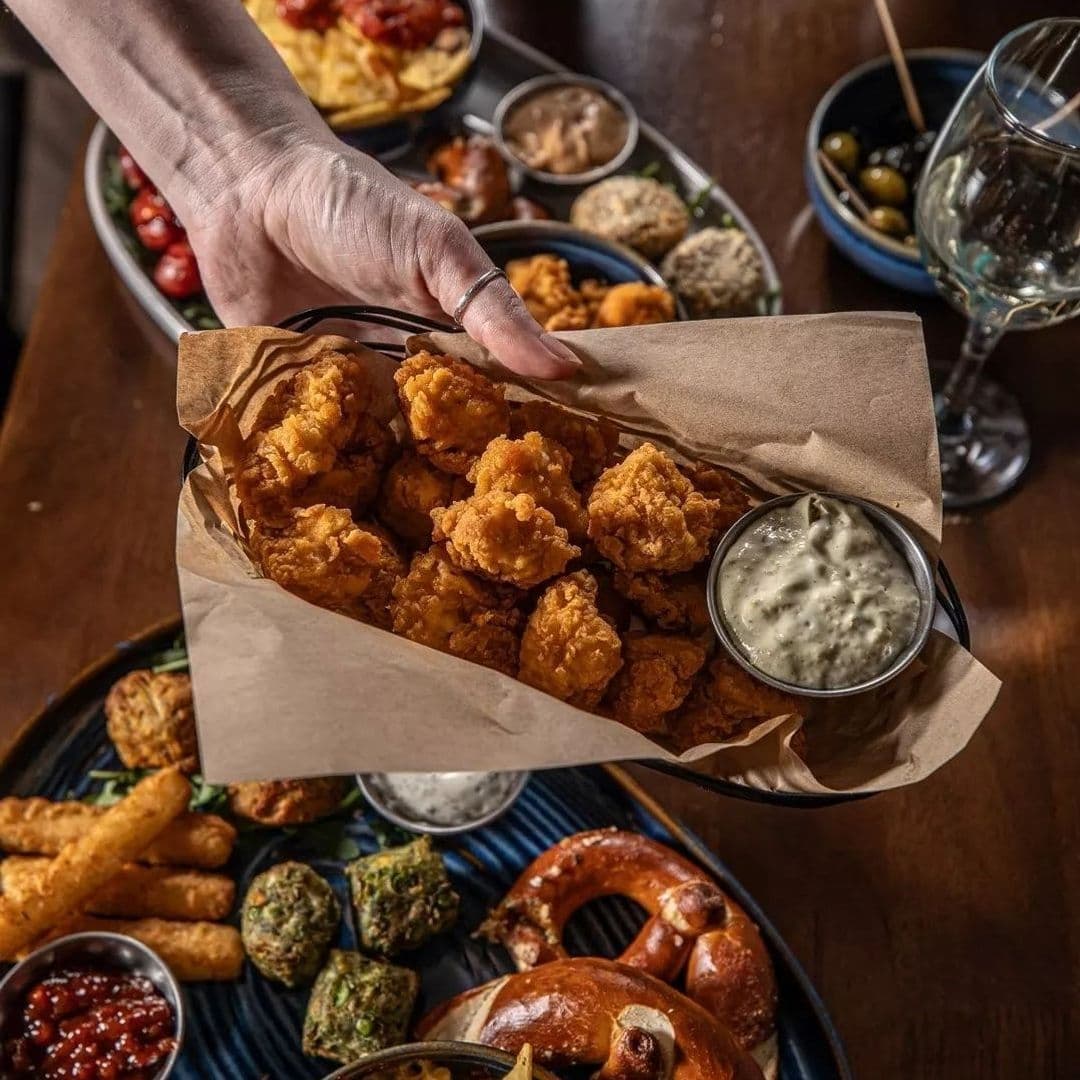 Alt text: "A person serving a basket of crispy chicken bites with dipping sauce at Parkside Tavern, surrounded by a variety of appetizers including pretzels, mozzarella sticks, and olives, with a glass of wine in the background."