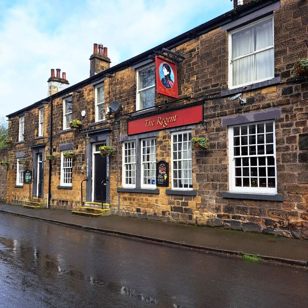 Exterior view of The Regent, a traditional stone-built pub with a red sign, white-framed windows, and hanging flower baskets on a wet street.