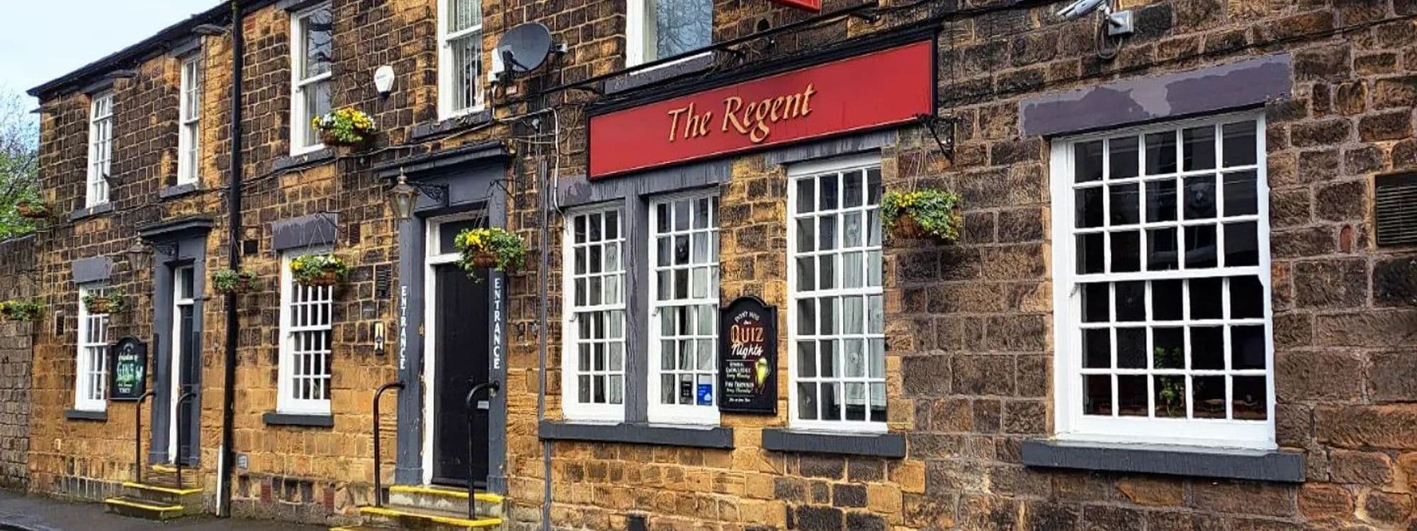 Exterior view of The Regent, a traditional stone-built pub with a red signboard, white-framed windows, and hanging flower baskets.
