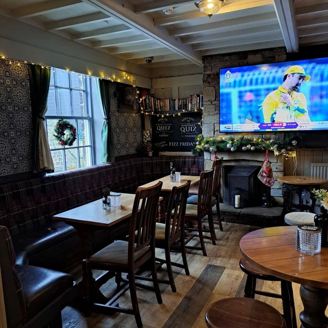 Cozy interior of Regent pub with wooden tables and chairs, patterned wallpaper, and a festive garland above a fireplace, with a large screen TV displaying a cricket match.