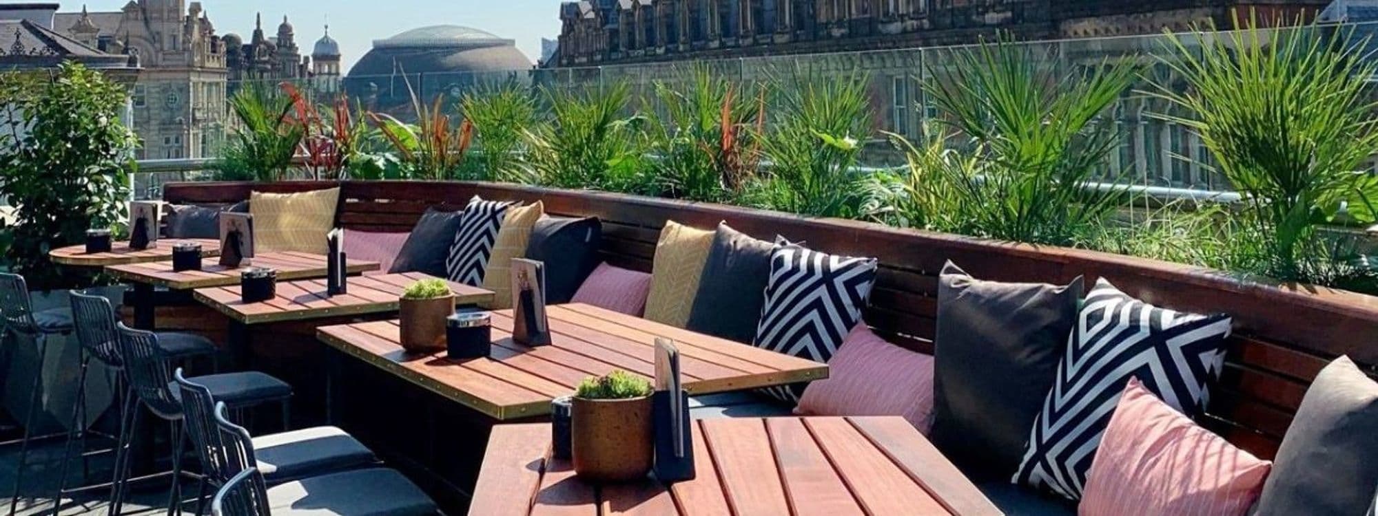 Outdoor seating area at The Alchemist Trinity with wooden tables, comfortable cushions, and lush green plants, offering a relaxing urban oasis for visitors.