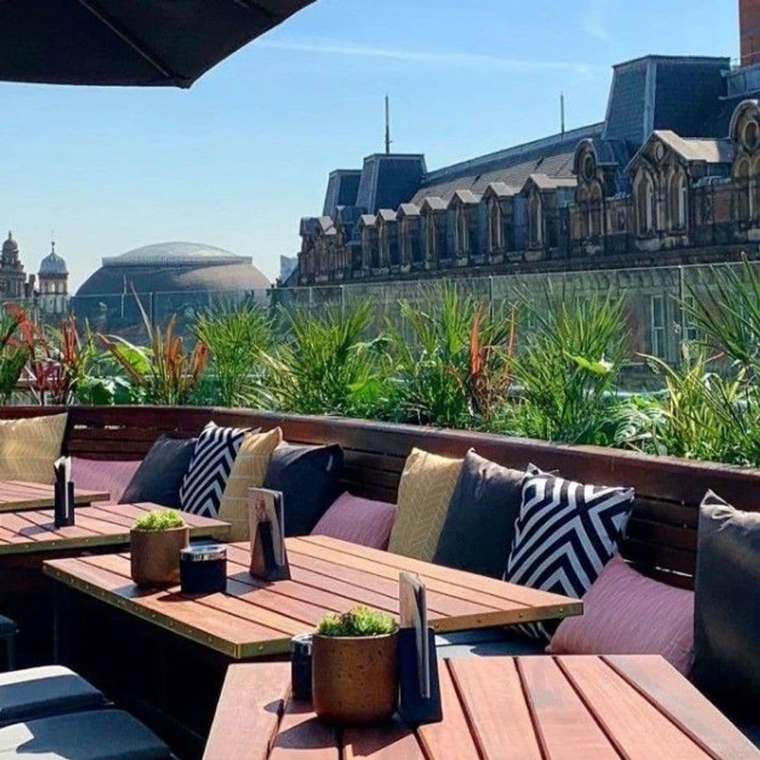 Outdoor seating area at The Alchemist Trinity with wooden tables, comfortable benches with cushions, and potted plants, overlooking historic architecture under a clear blue sky.