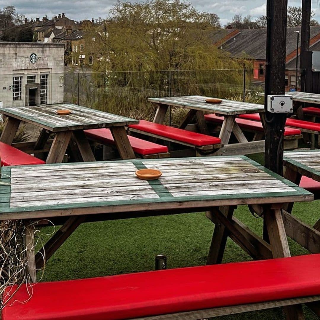 Outdoor seating area at The Woods with wooden picnic tables and red benches on a green artificial turf, overlooking urban surroundings.