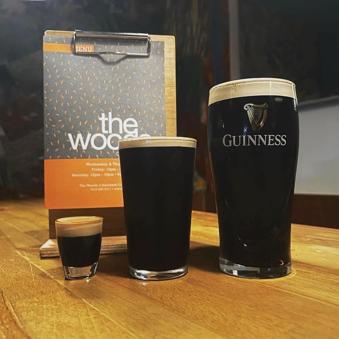 A pint of Guinness beer and a small shot glass of dark liquor on a wooden table, with a menu clipboard labeled 'The Woods' in the background.