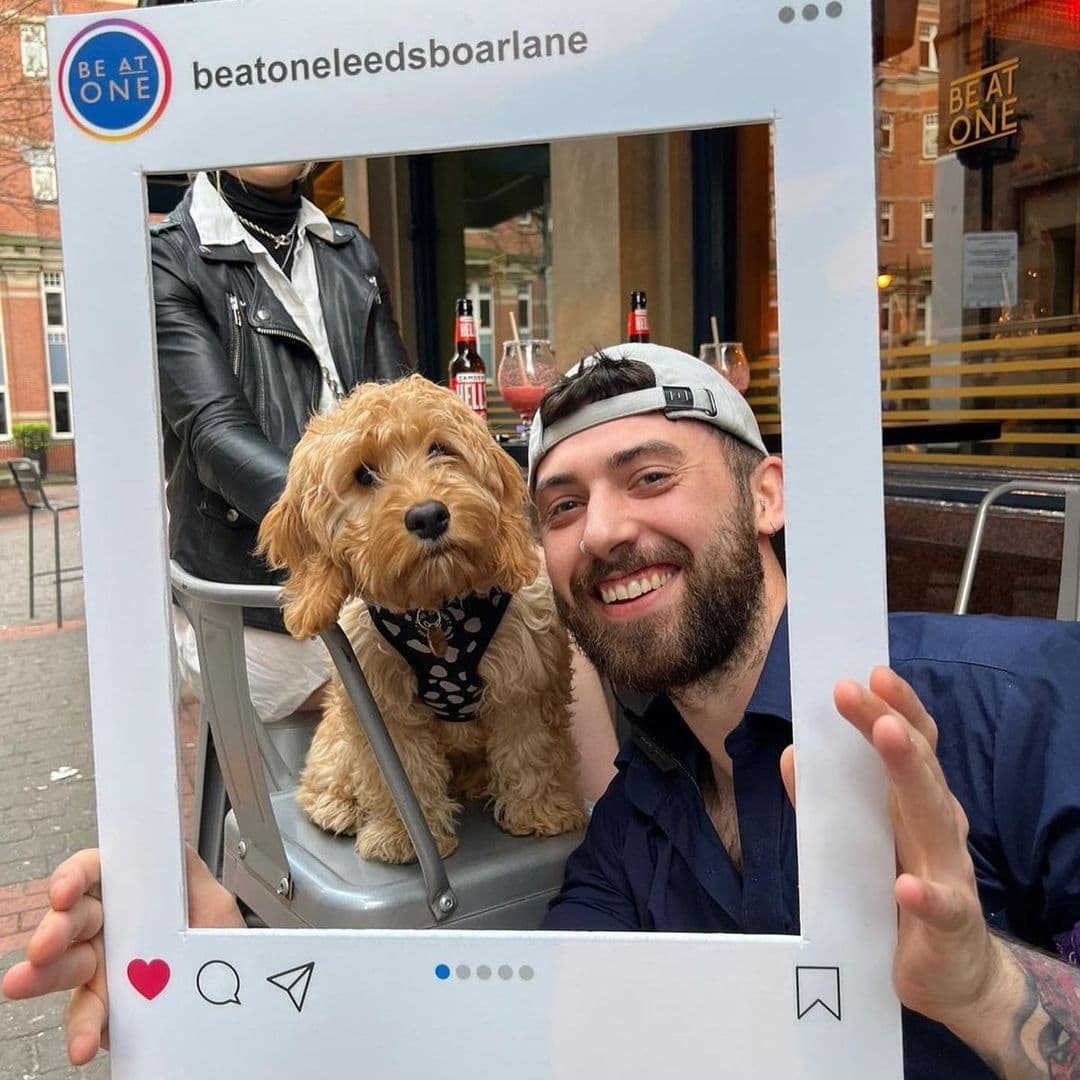 Smiling man with a beard and backward cap posing with a cute dog in a bandana through a large frame, with Be At One – Boar Lane signage in the background.