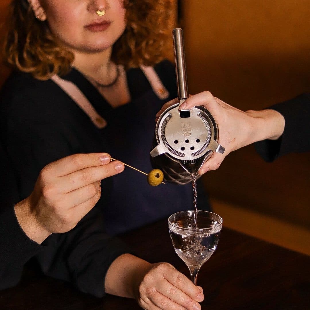 Bartender at Below Stairs pouring a cocktail through a strainer into a glass, with an olive on a pick.