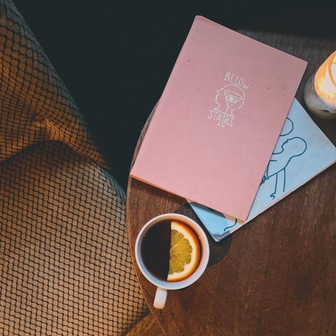A cozy setting at Below Stairs featuring a cup of tea with a lemon slice on a wooden table, accompanied by a pink menu with the establishment's name and a sketchbook with a blue cover.