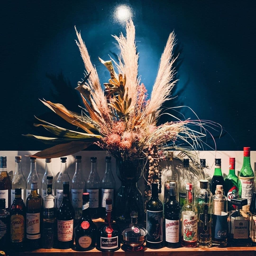 A variety of liquor bottles on a bar shelf at Below Stairs with an elegant arrangement of pampas grass and dried foliage illuminated by mood lighting.