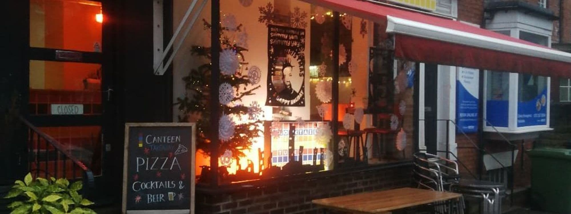 Exterior view of Canteen with warm lighting, decorated with white snowflake ornaments and a Christmas tree, featuring a sign advertising pizza, cocktails, and beer.