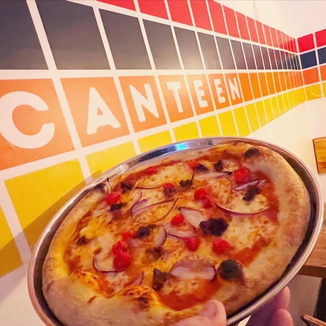 Freshly baked pizza with toppings being served at Canteen, with the restaurant's colorful wall featuring the name 'CANTEEN' in large letters in the background.