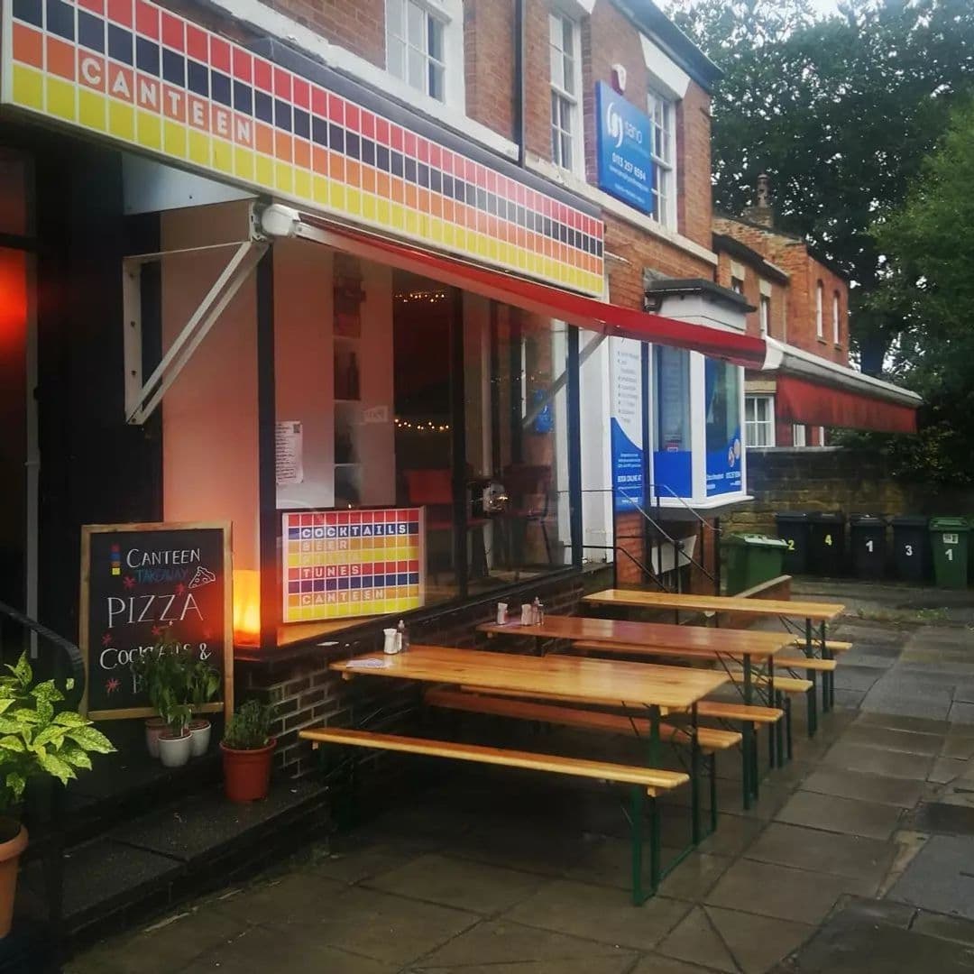 Exterior view of Canteen, featuring a colorful tiled facade with signage, large windows, outdoor wooden benches, and potted plants on a paved sidewalk.