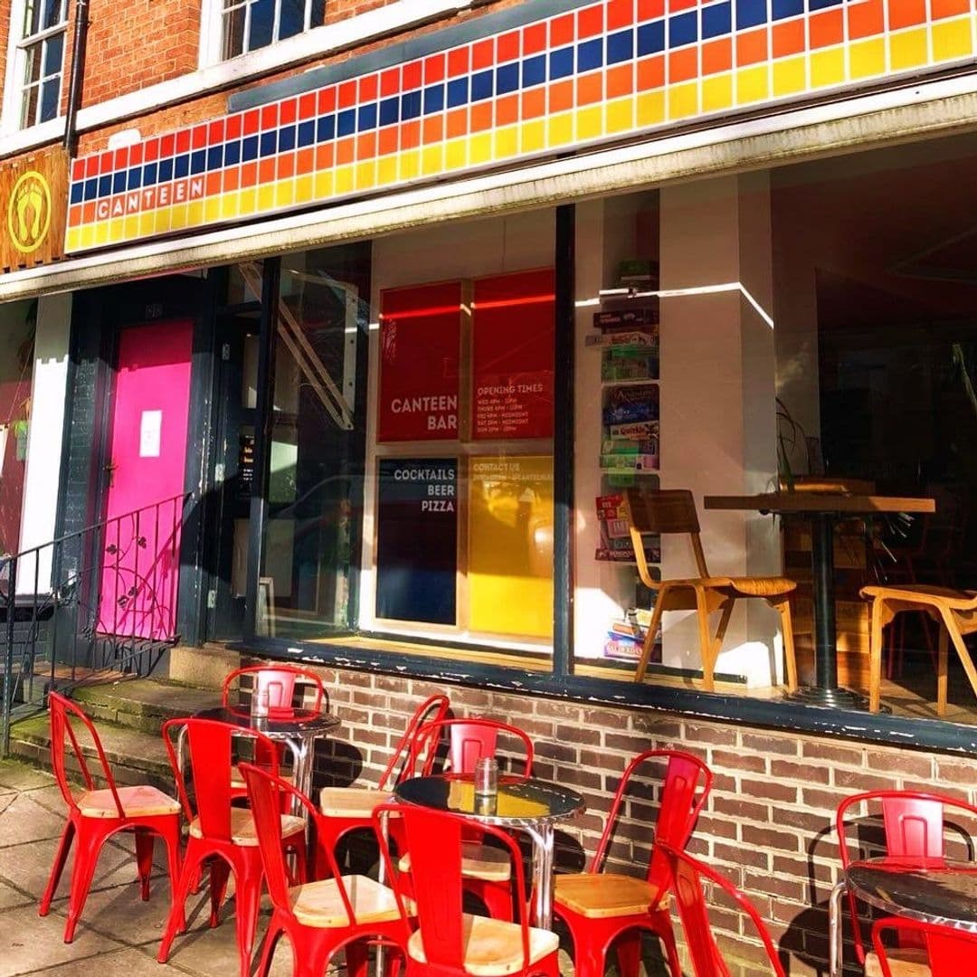 Exterior view of Canteen with colorful tile facade, red outdoor chairs, and wooden tables under sunny skies.
