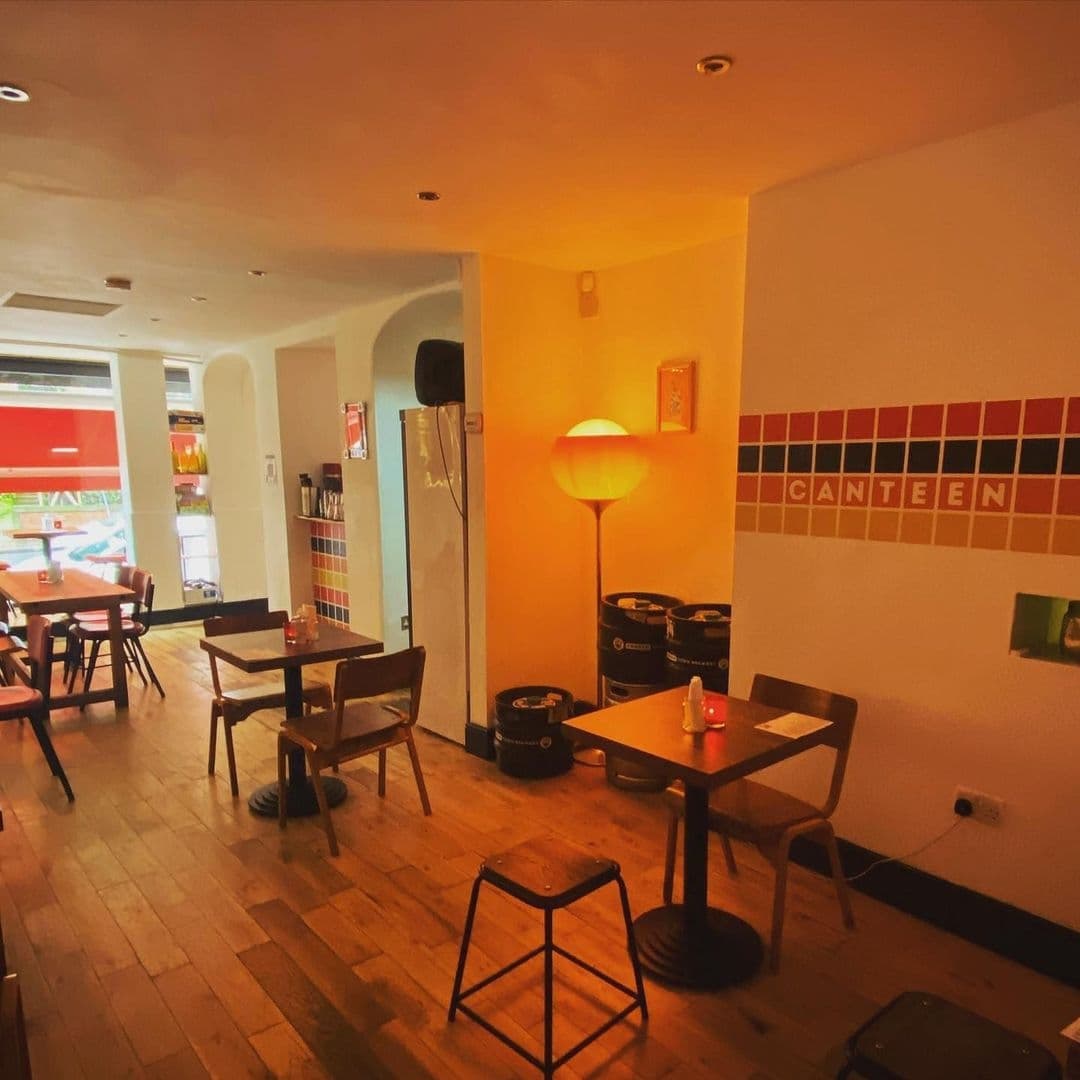 Interior of Canteen featuring wooden tables and chairs, warm lighting from a floor lamp, and decorative barrels with a tiled wall sign spelling out 'CANTEEN'.