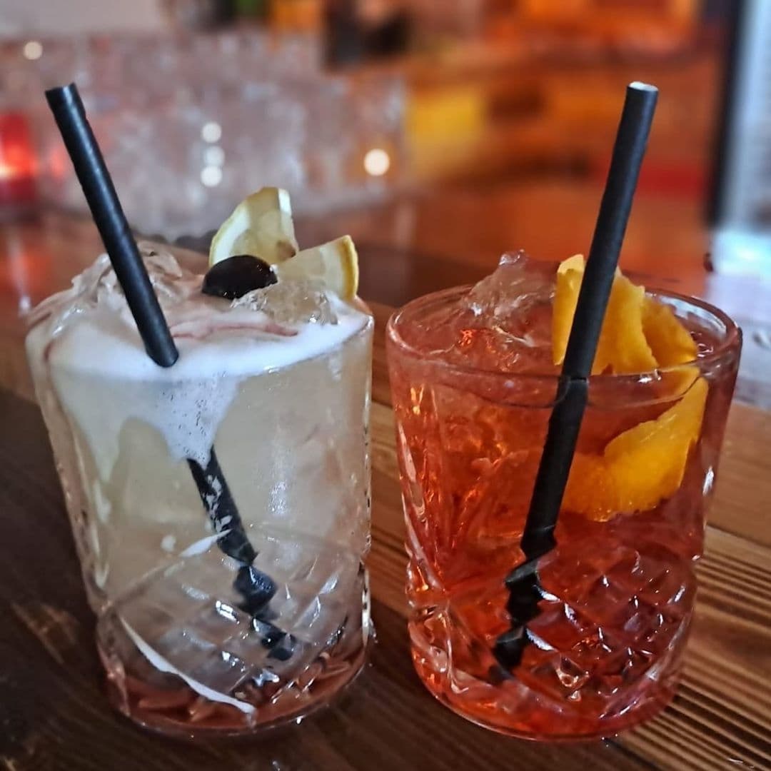 Two specialty cocktails on a bar counter at Canteen, with one featuring a frothy top and garnished with a lemon twist and a berry, and the other with an orange peel garnish, both with black straws.