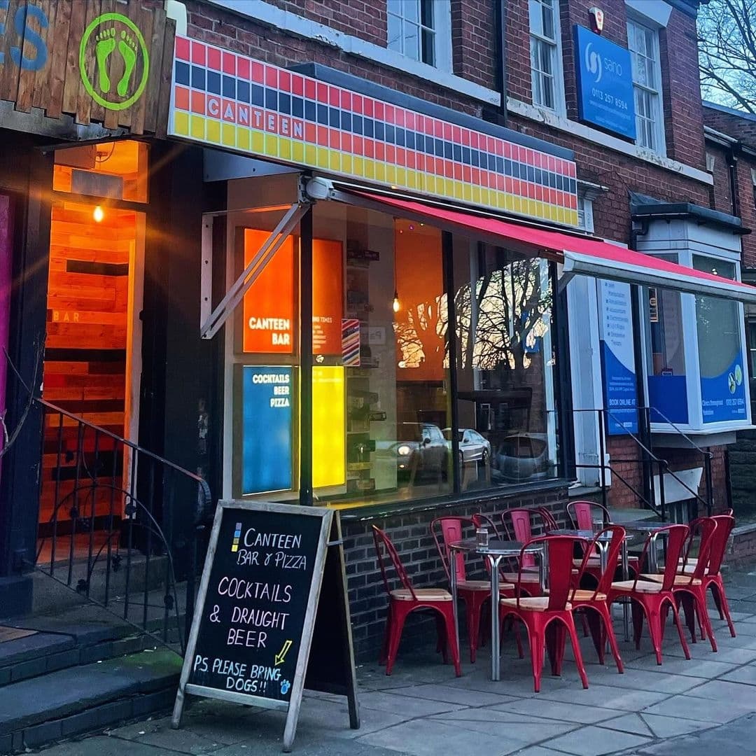 Exterior view of Canteen bar and pizza restaurant at dusk with colorful facade, outdoor red seating, and a welcoming signboard advertising cocktails, draught beer, and an invitation for dogs.