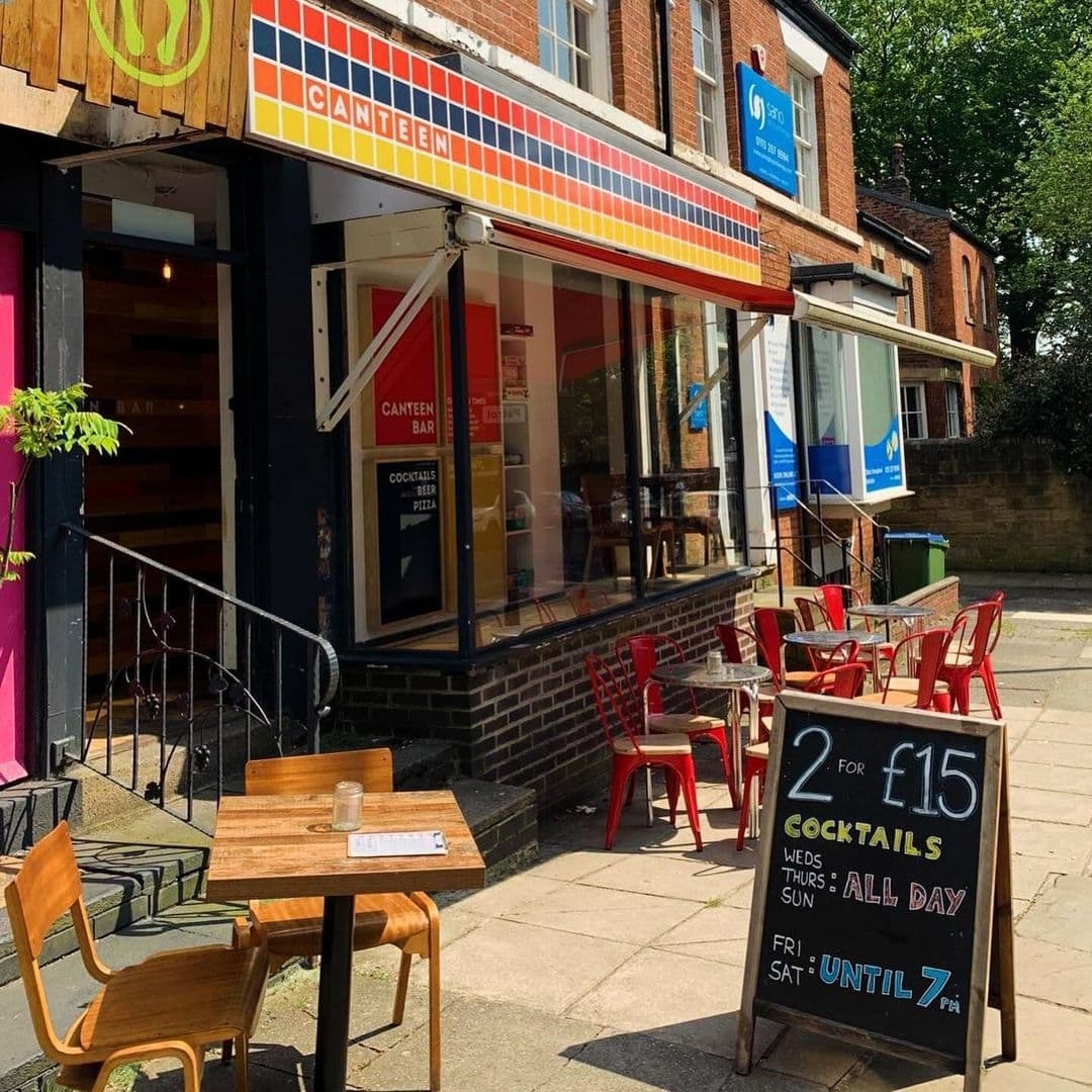 Exterior view of Canteen, featuring a vibrant storefront with a colorful striped awning, outdoor seating with wooden tables and red chairs, and a chalkboard sign advertising cocktail deals.