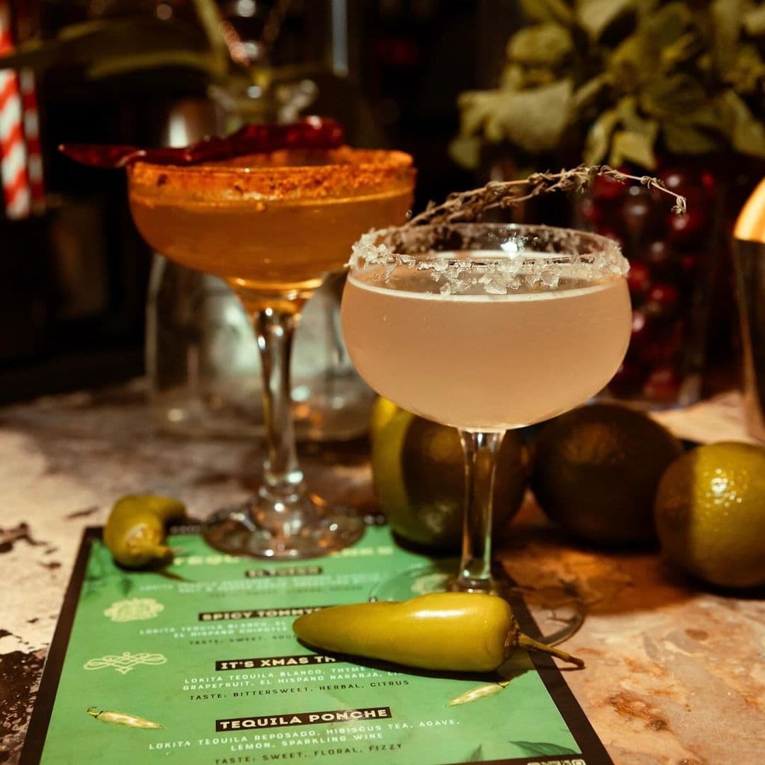 Two specialty cocktails on a bar menu at Distrikt, with a rustic ambiance and garnishes like a chili pepper and a sprig of herbs.