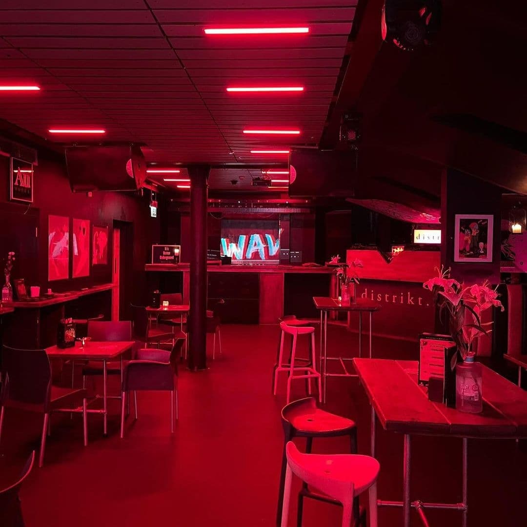 Interior of Distrikt, featuring red neon lighting, a bar area with stools, and a stage with a large screen displaying 'WAV'. Tables with chairs are set around the room under a ceiling with linear lighting fixtures.