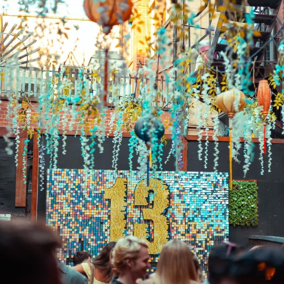 Vibrant outdoor atmosphere at Distrikt with colorful hanging decorations, a mosaic wall featuring the number 13, and people socializing in the background.