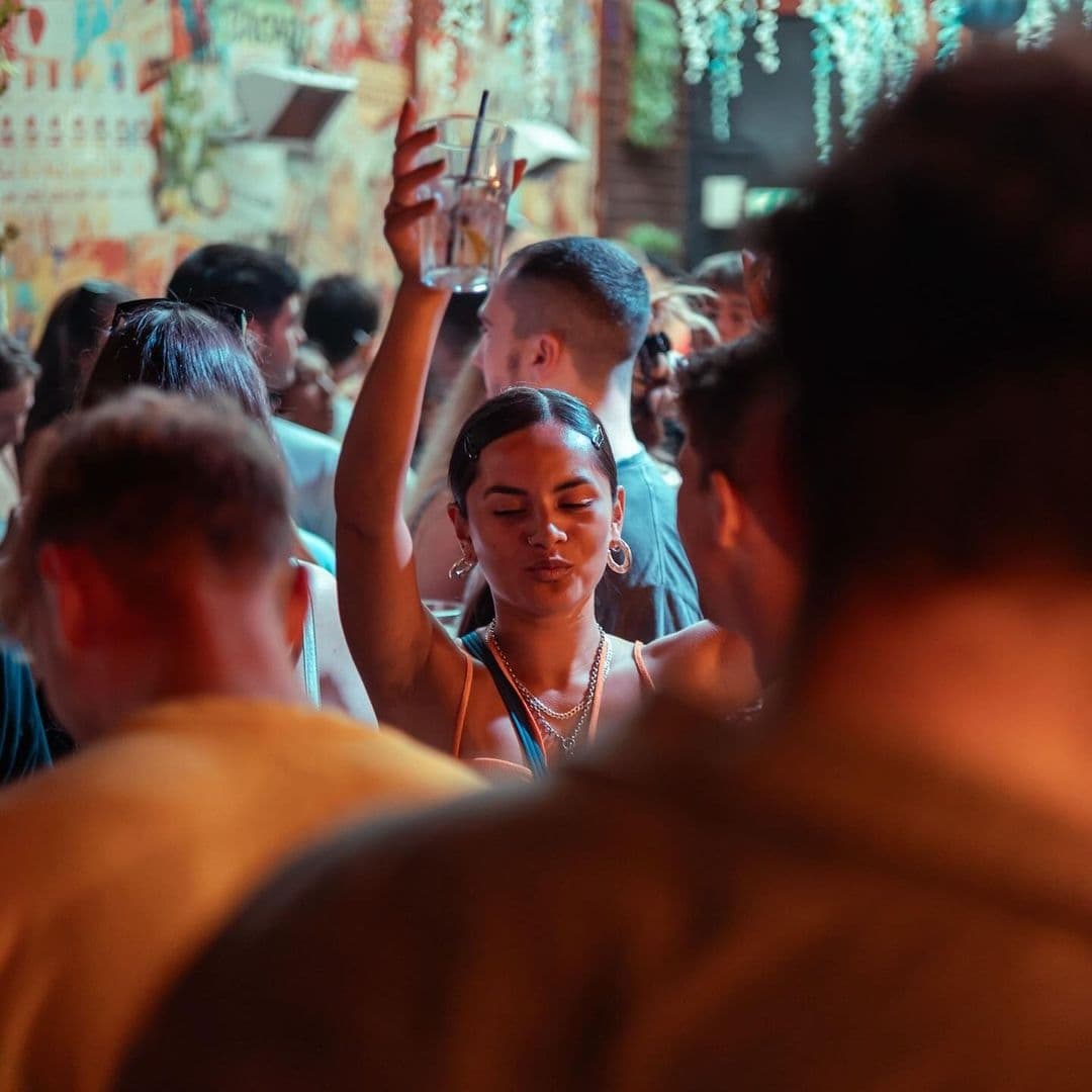 Vibrant nightlife at Distrikt with a young woman enjoying a drink and dancing among a crowd of people, with colorful lights and murals in the background.