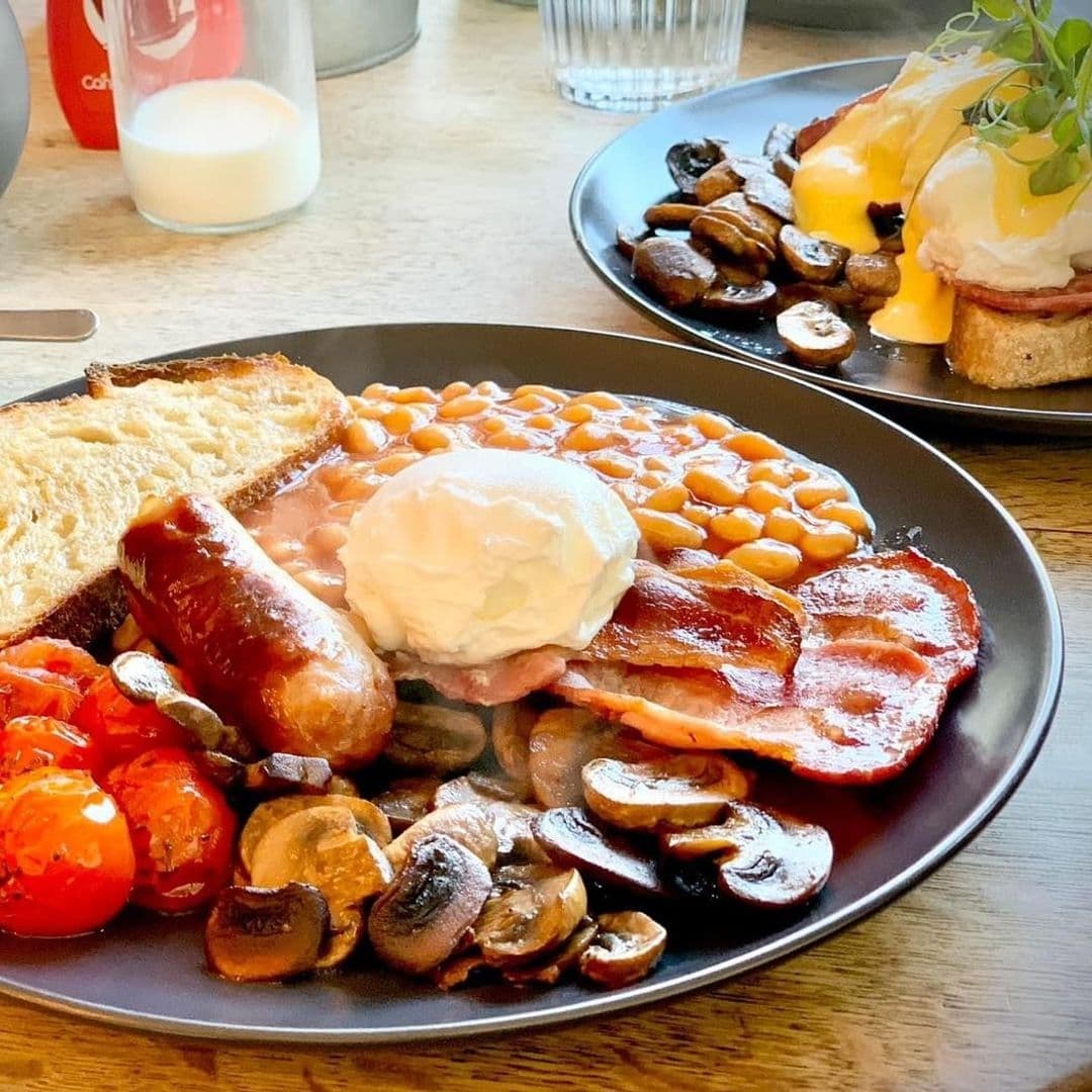 Traditional English breakfast served at Gray's featuring eggs, bacon, sausages, baked beans, grilled tomatoes, mushrooms, and toast on a black plate, with a glass of milk and condiments in the background.