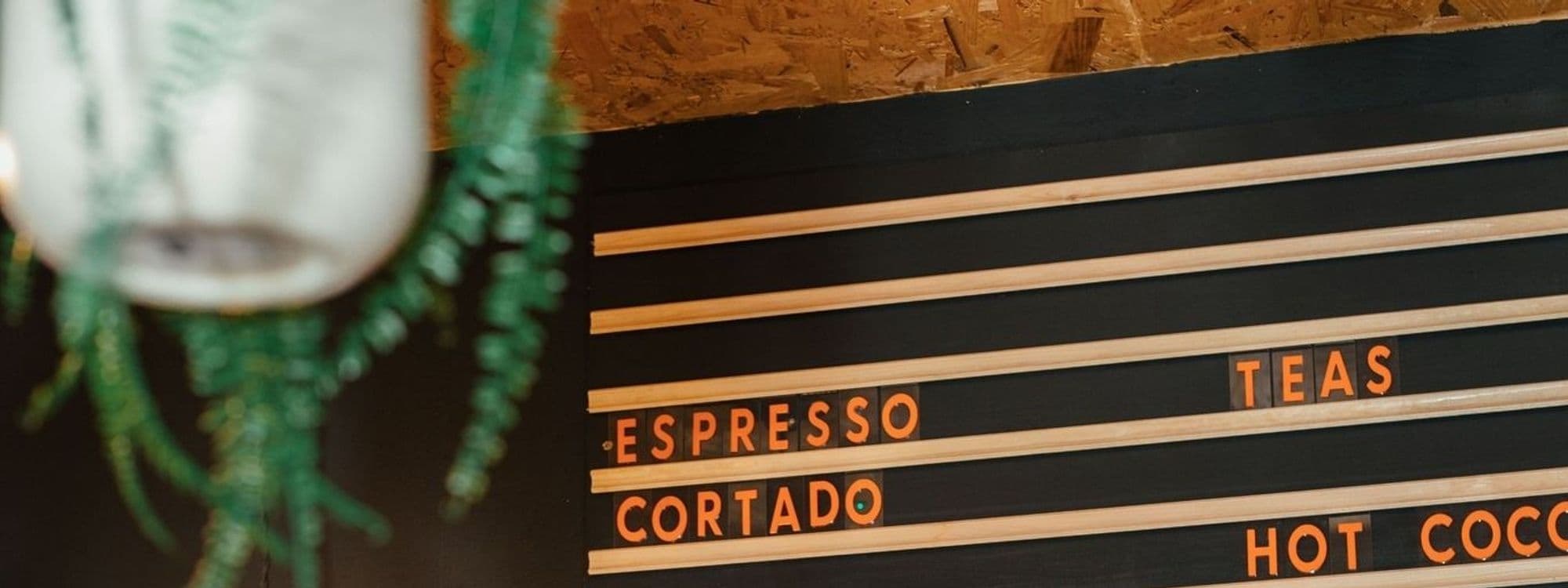 Interior detail of Livett's Coffee Shop featuring a menu board with beverage options such as espresso, cortado, teas, and hot cocoa, with a blurred plant in the foreground.