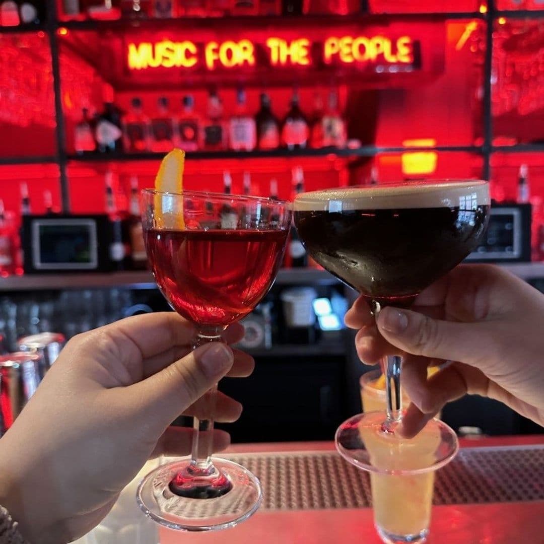 Two hands holding cocktails with a red neon sign reading 'MUSIC FOR THE PEOPLE' in the background at MOJO.