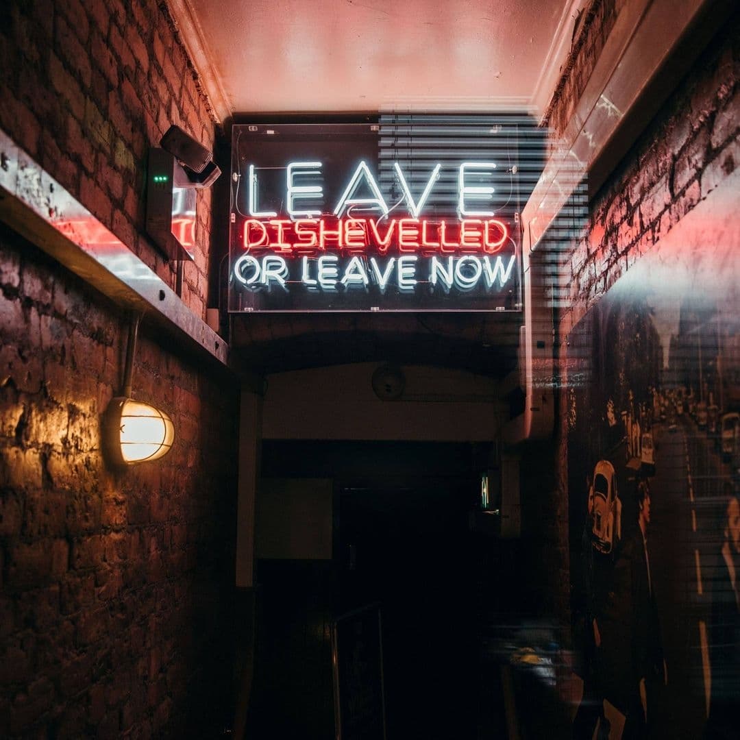 Neon sign at MOJO with the quirky message "Leave Dishevelled or Leave Now" above a brick-walled corridor, evoking the venue's edgy and playful atmosphere.