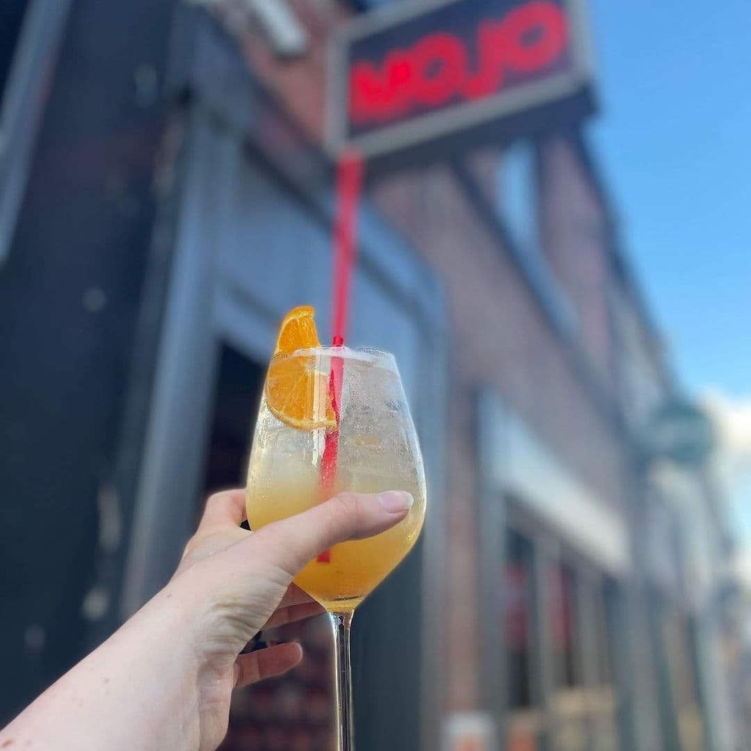 Hand holding a cocktail with an orange slice garnish in front of the MOJO sign.