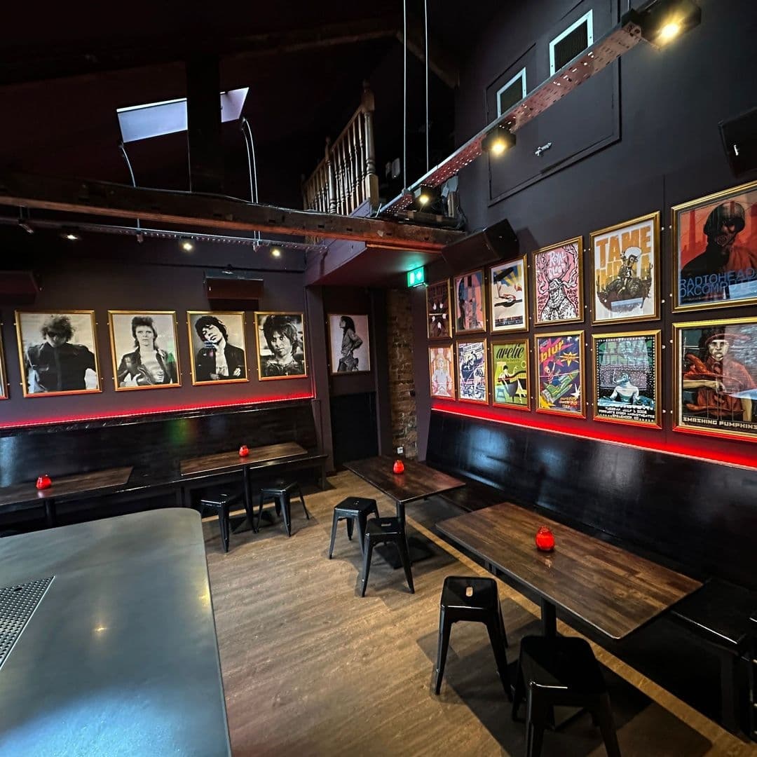 Interior of MOJO featuring a cozy setting with black stools and tables, red accent lighting, and walls adorned with framed pictures of iconic music and pop culture figures. A balcony overlooks the space, enhancing the venue's intimate atmosphere.