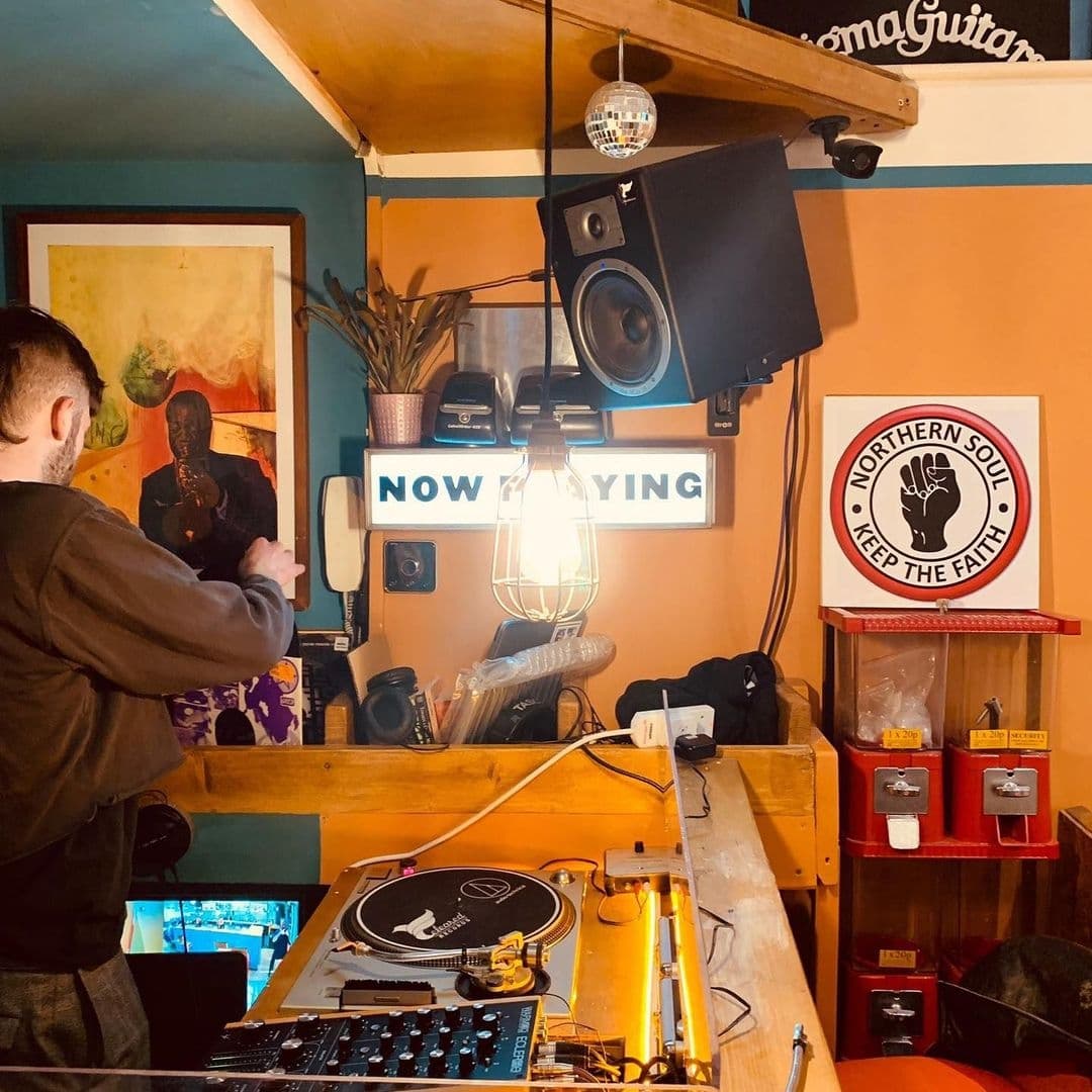 A DJ booth at Melodie 71 featuring a turntable, mixer, and a 'NOW PLAYING' sign, with a person selecting records, a disco ball, and music-themed decor.