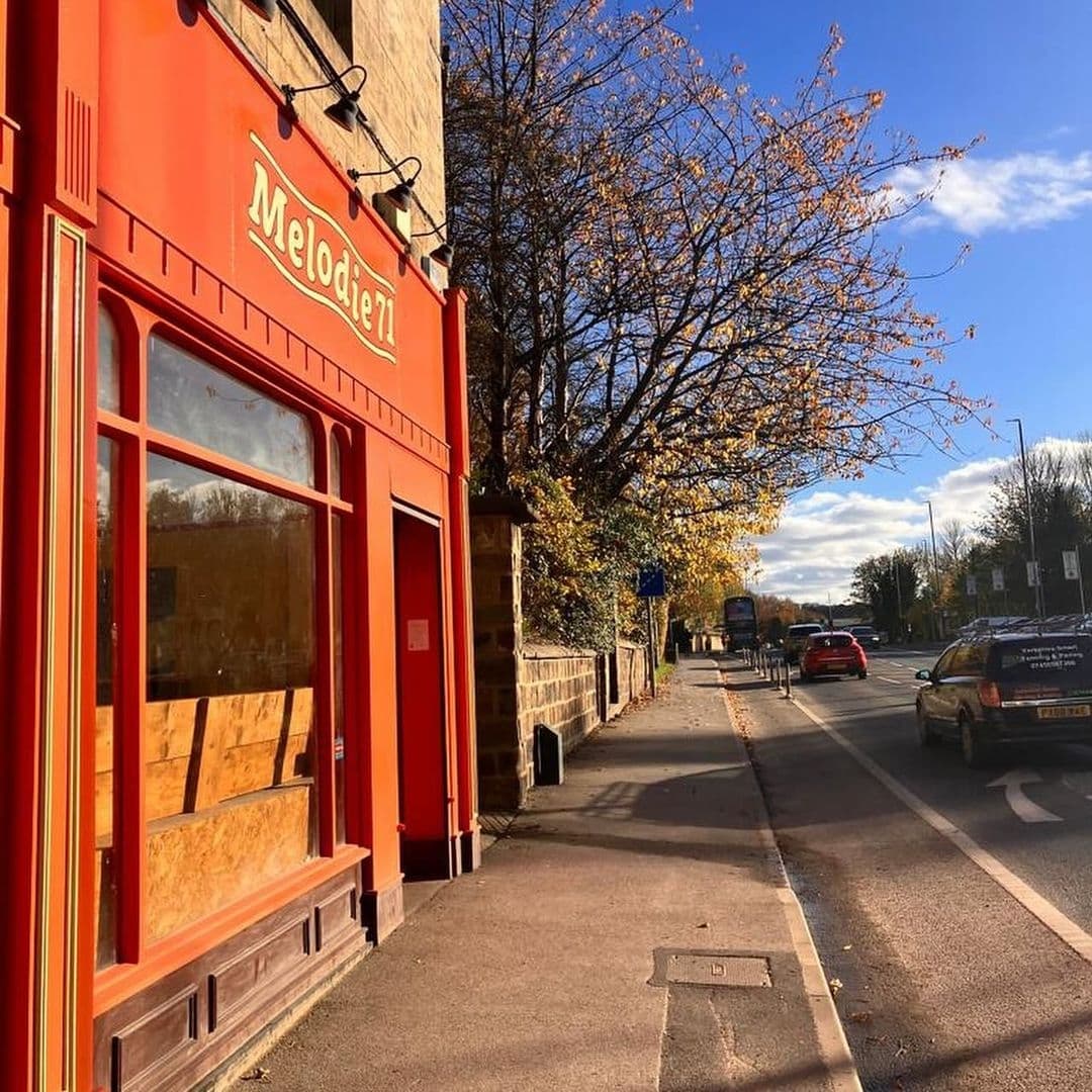 Exterior view of Melodie 71 on a sunny day, showcasing the vibrant red facade with large windows, set against a street with autumn trees and a clear blue sky.