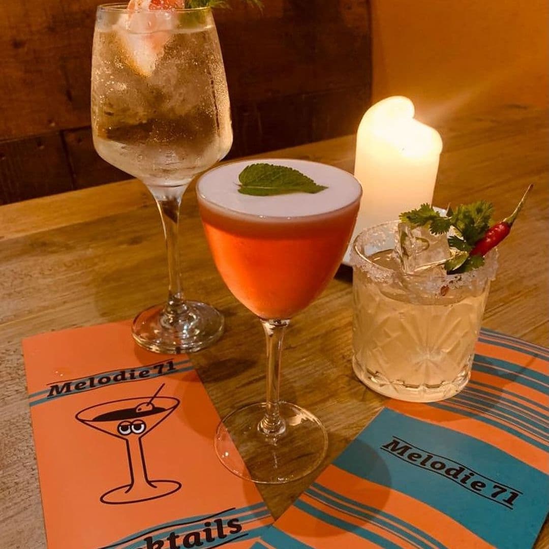 Three assorted cocktails on a table at Melodie 71, with a menu featuring the establishment's name.